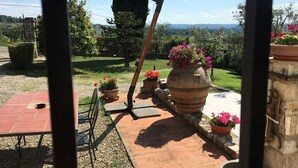 Une terrasse extérieure avec une table et des chaises, entourée de plantes en pot et d’un grand vase en pierre.