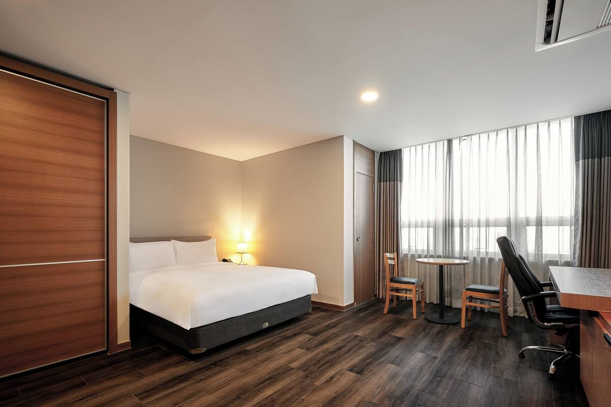 Habitación de hotel con una cama, un escritorio, dos sillas, una lámpara y una ventana con cortinas.