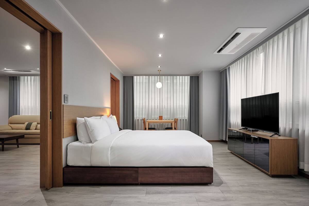 Una habitación de hotel moderna con una cama grande, un televisor, una mesita de madera y un sofá al fondo.