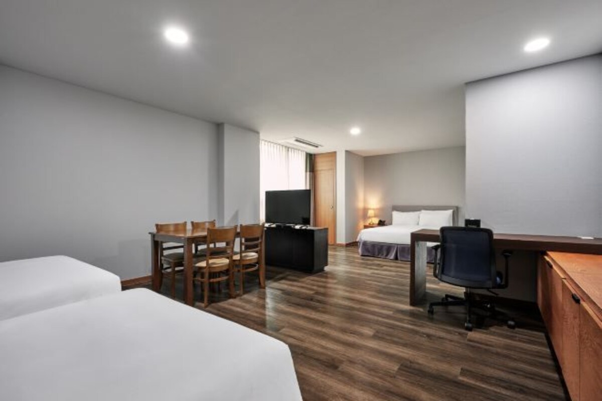 Habitación de hotel con dos camas individuales, un escritorio con una silla, un televisor y una pequeña zona de estar con sillones.