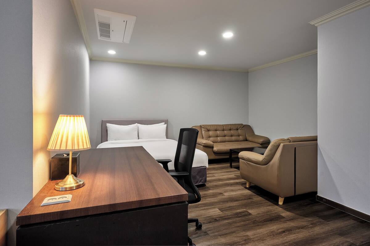 Una habitación de hotel con un escritorio de madera, una lámpara, una cama con ropa blanca, un sillón de cuero y un sofá.