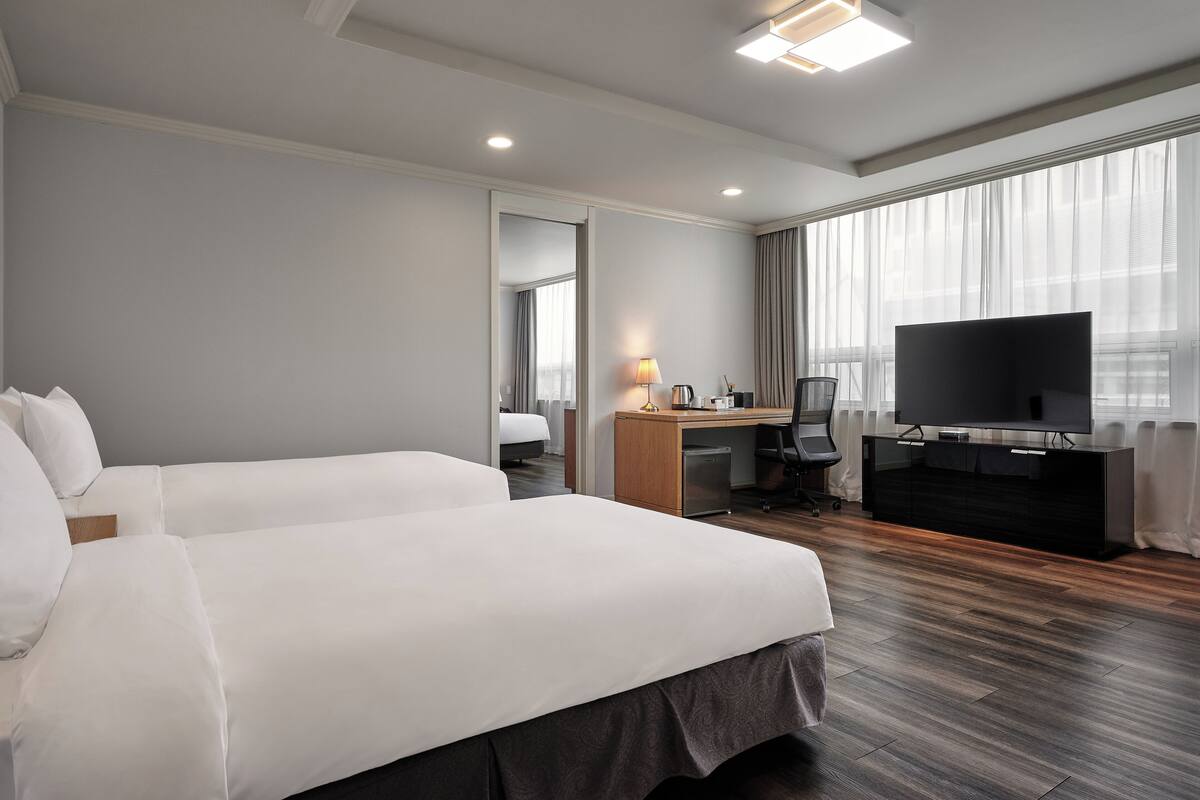 Habitación de hotel con dos camas, un escritorio, un televisor y un ventanal con cortinas.
