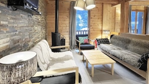 A cozy cabin interior with a stone wall, wooden furniture, a fireplace, and a view of the outdoors.