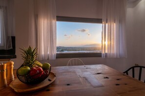 Un comedor con una mesa de madera, una cesta de frutas y vista al atardecer a través de una ventana con cortinas livianas.