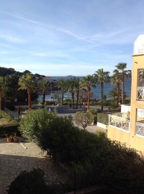 A coastal view with palm trees, a paved area, and a building with a balcony. - PORTISSOL T2: Patios with ocean views, 100 meters from the beach (Sanary-sur-Mer)