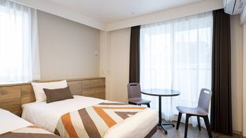 A hotel room with two beds, a small table, a chair, and large windows with curtains.