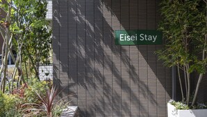 Un letrero que dice 'Eisei Stay' en la fachada de un edificio de diseño moderno, rodeado de follaje y plantas.