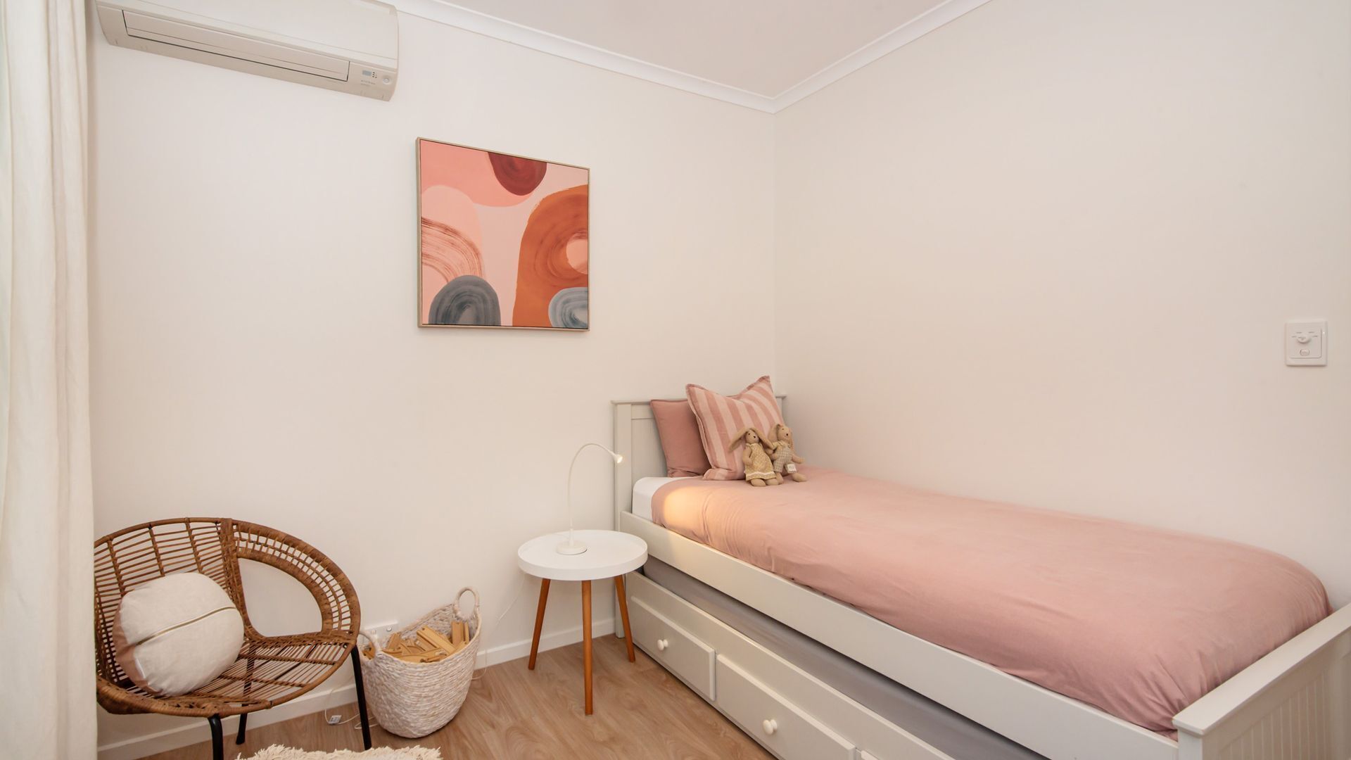 Un dormitorio con una cama individual, una silla de mimbre, una mesita auxiliar y un aire acondicionado de pared.