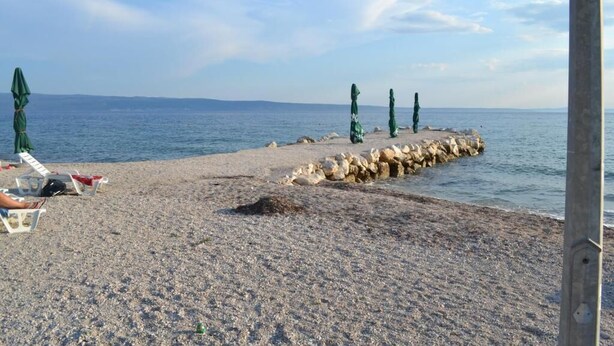 Ein Kiesstrand mit einem steinernen Wellenbrecher, einigen Sonnenschirmen und Liegestühlen.