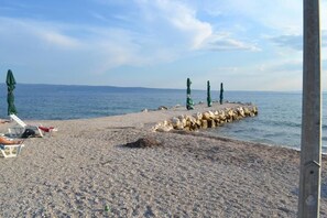 A pebbled beach with a rocky breakwater, a few umbrellas, and lounge chairs. - Apartments Lucia (Podstrana)