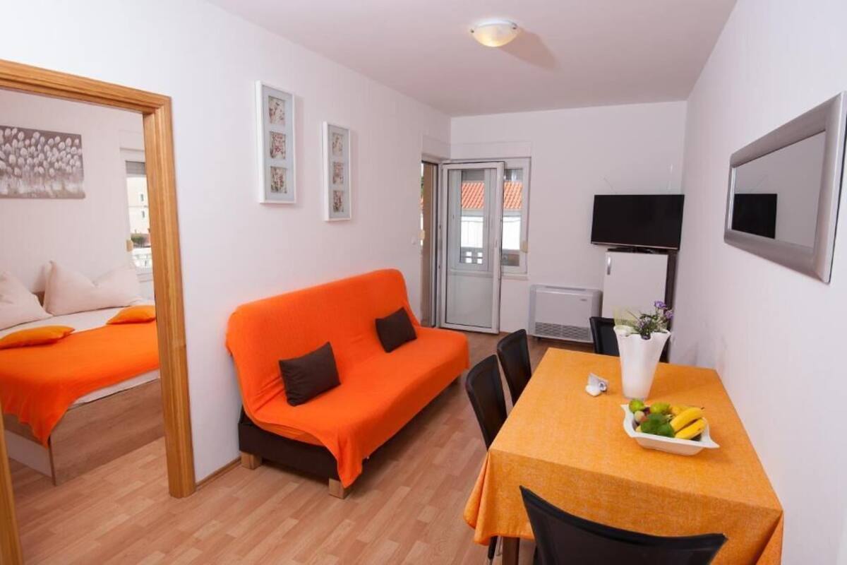 A compact room with a bed, an orange sofa, a dining table with a fruit bowl, a TV, and a mirror.
