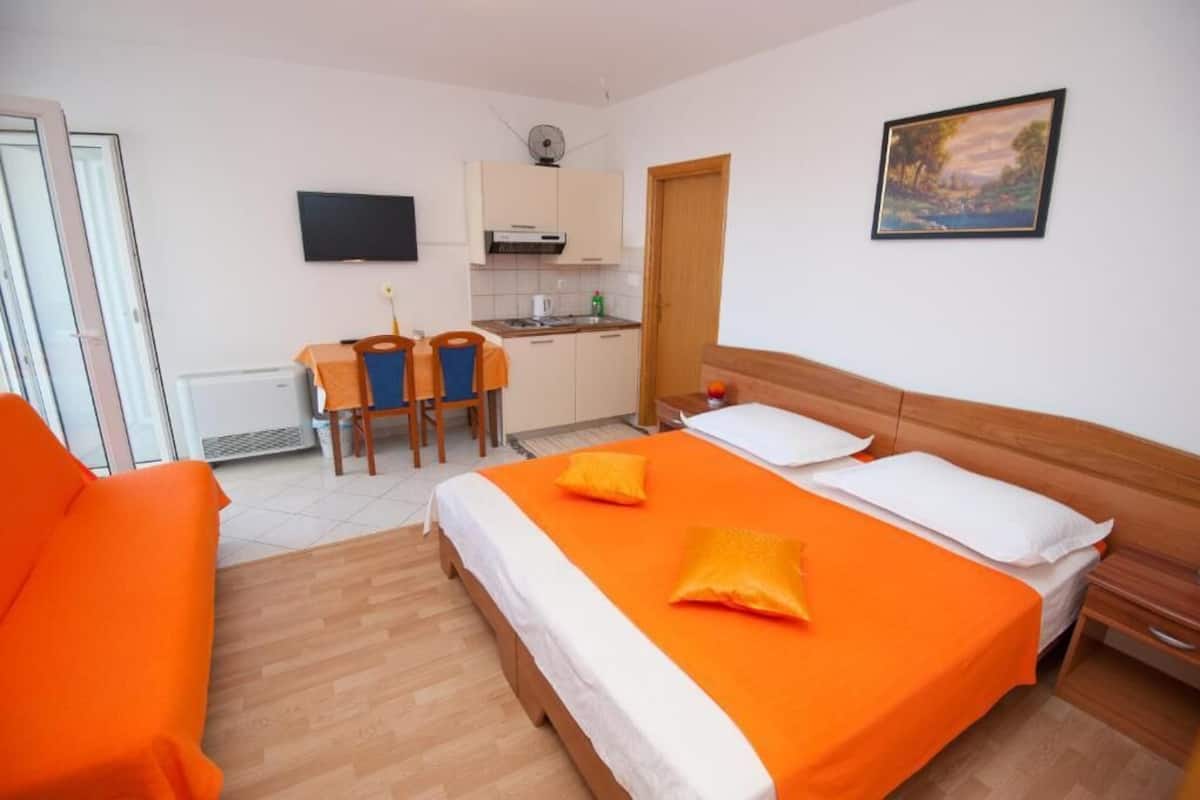 A hotel room with a bed, a sofa, a small kitchen area, and a TV.