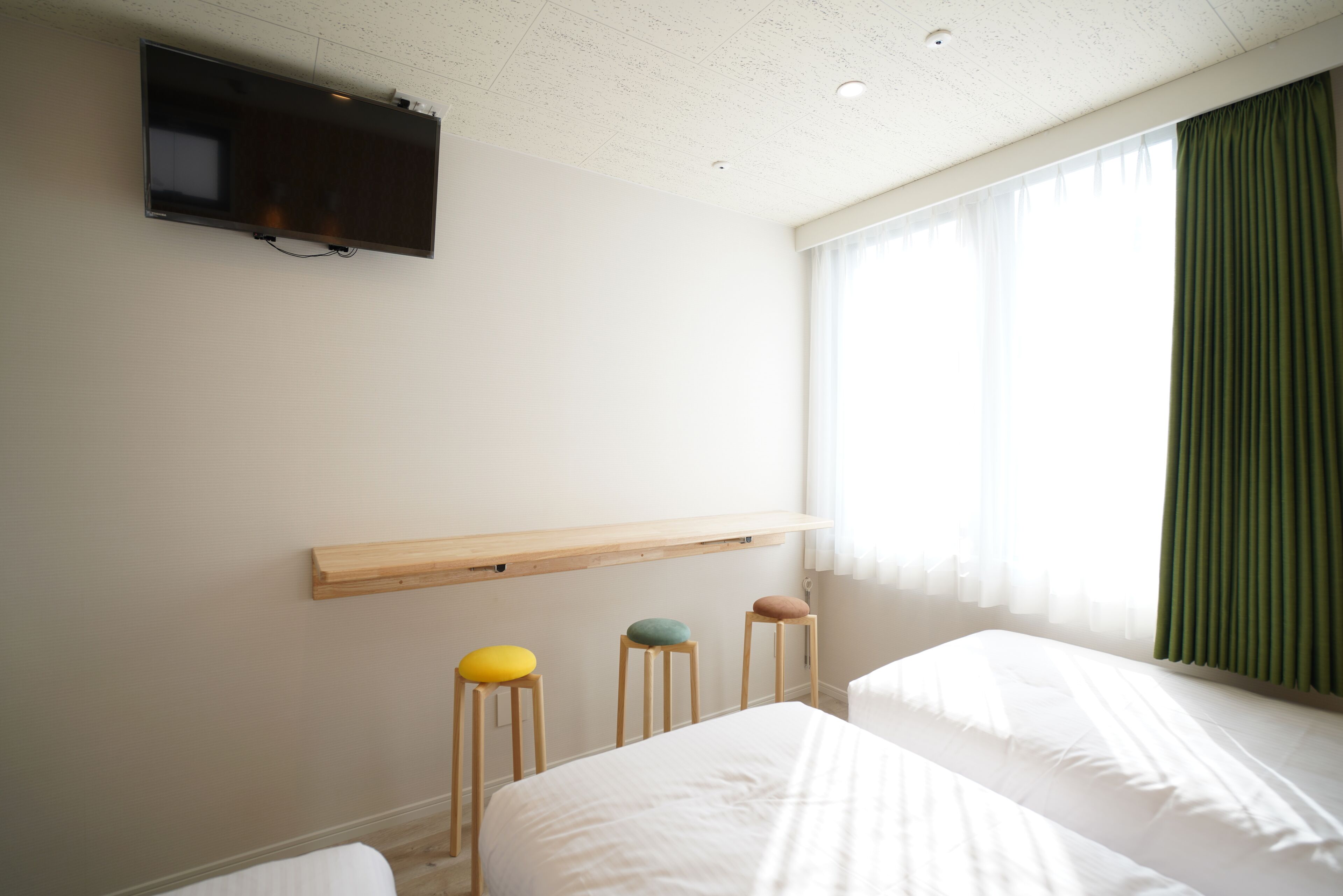 Quarto de hotel com televisão de tela plana na parede, prateleira de madeira sobre a cama e três banquinhos coloridos.