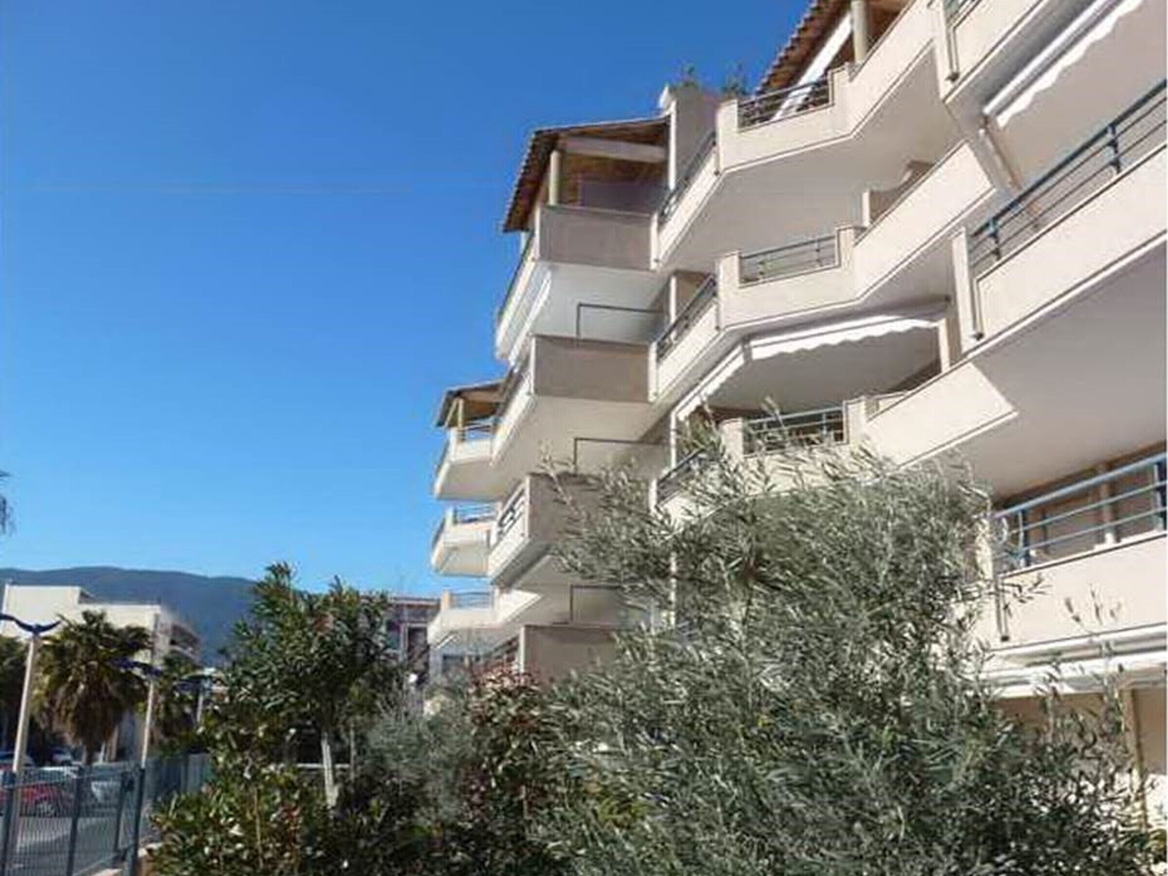 A multi-story residential building with balconies, surrounded by greenery and a clear sky.