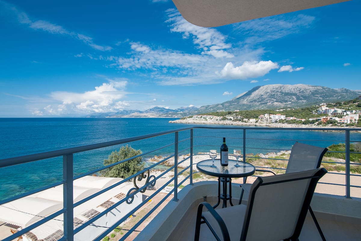 A balcony with a table and chairs overlooking a coastal view with mountains in the background.