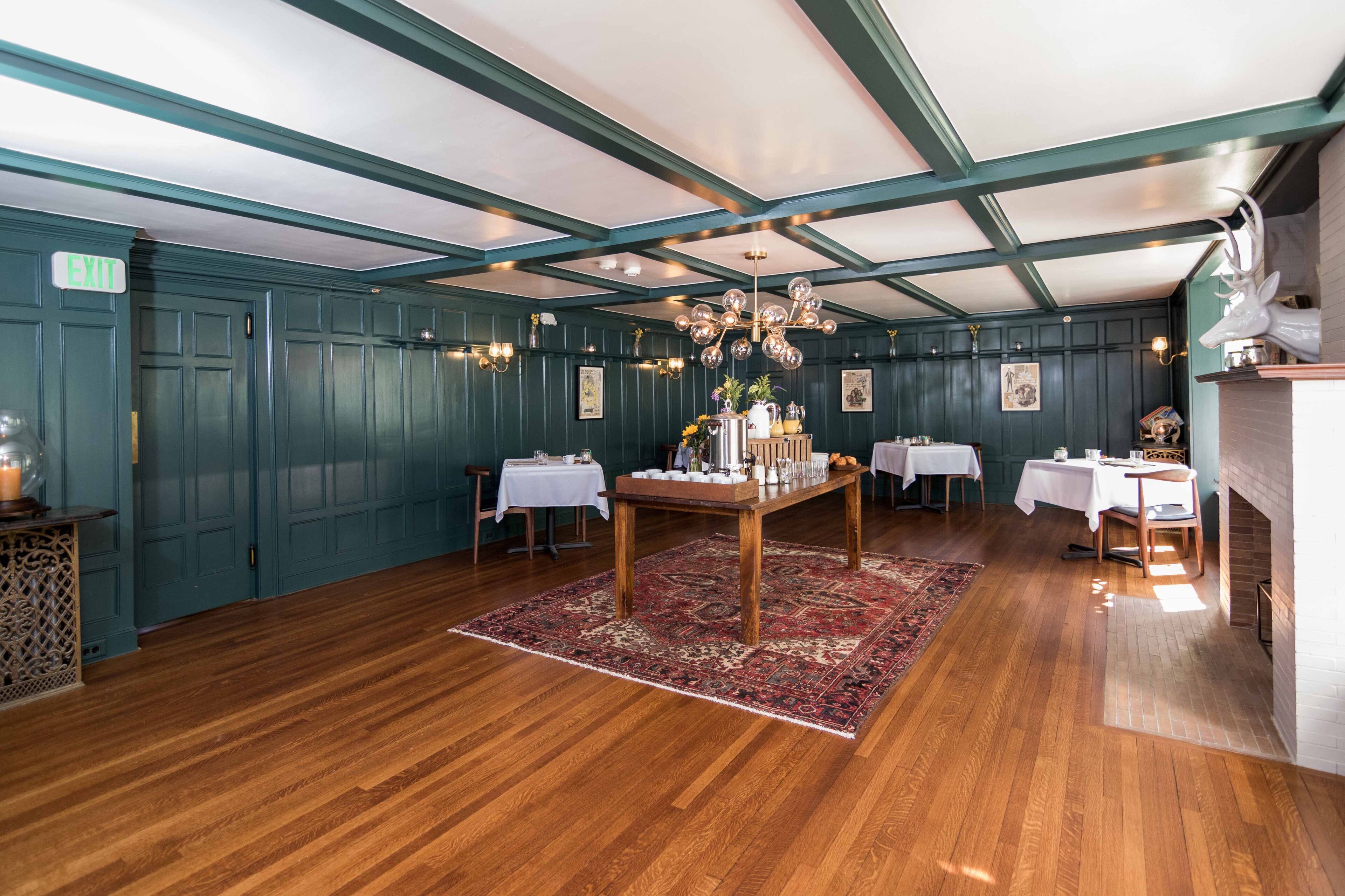 Una habitación con pisos de madera, paredes color turquesa y un candelabro. Hay mesas con manteles blancos y una chimenea.