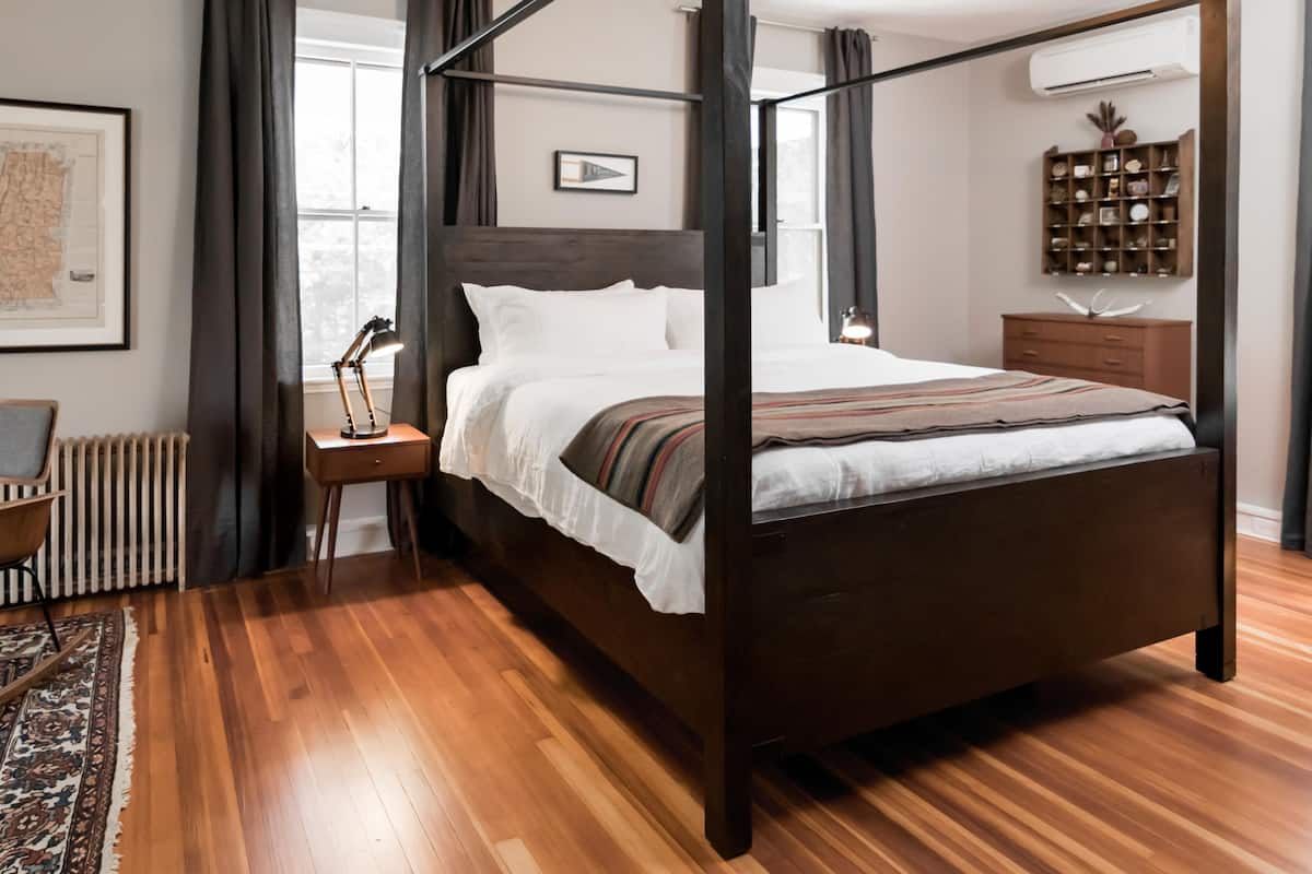 A four-poster bed in a well-lit room with a wooden floor, a nightstand with a lamp, a radiator, a bookshelf, and a framed picture on the wall.