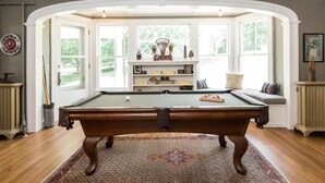 A pool table in a room with a patterned rug, a shelf with decorative items, and a window with a view of greenery. - Granville House (Housatonic)
