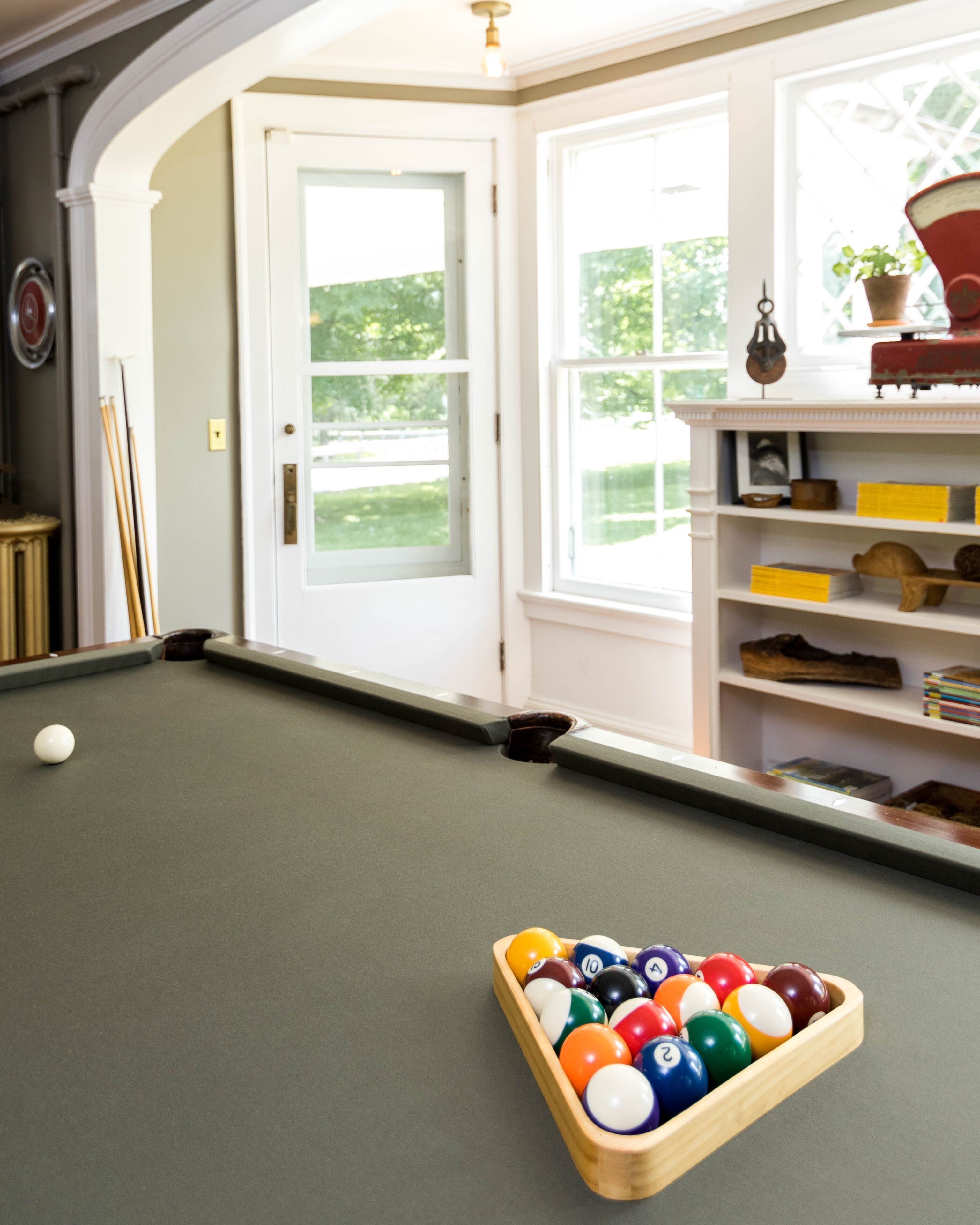 A pool table with a wooden ball rack in the foreground, a door leading to a garden, and a shelf with various items.