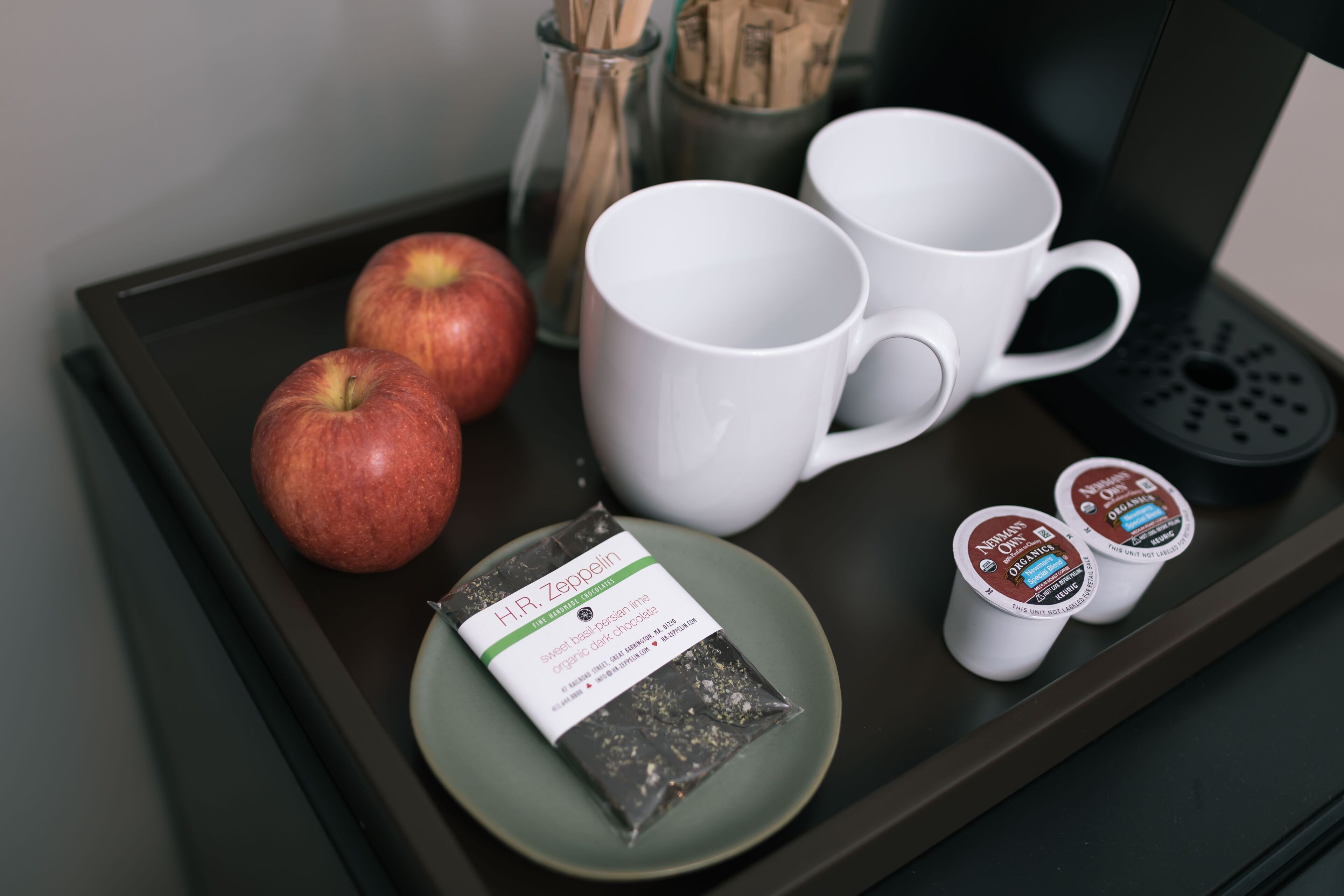 A tray with two apples, two white mugs, and two small containers of organic dark chocolate.
