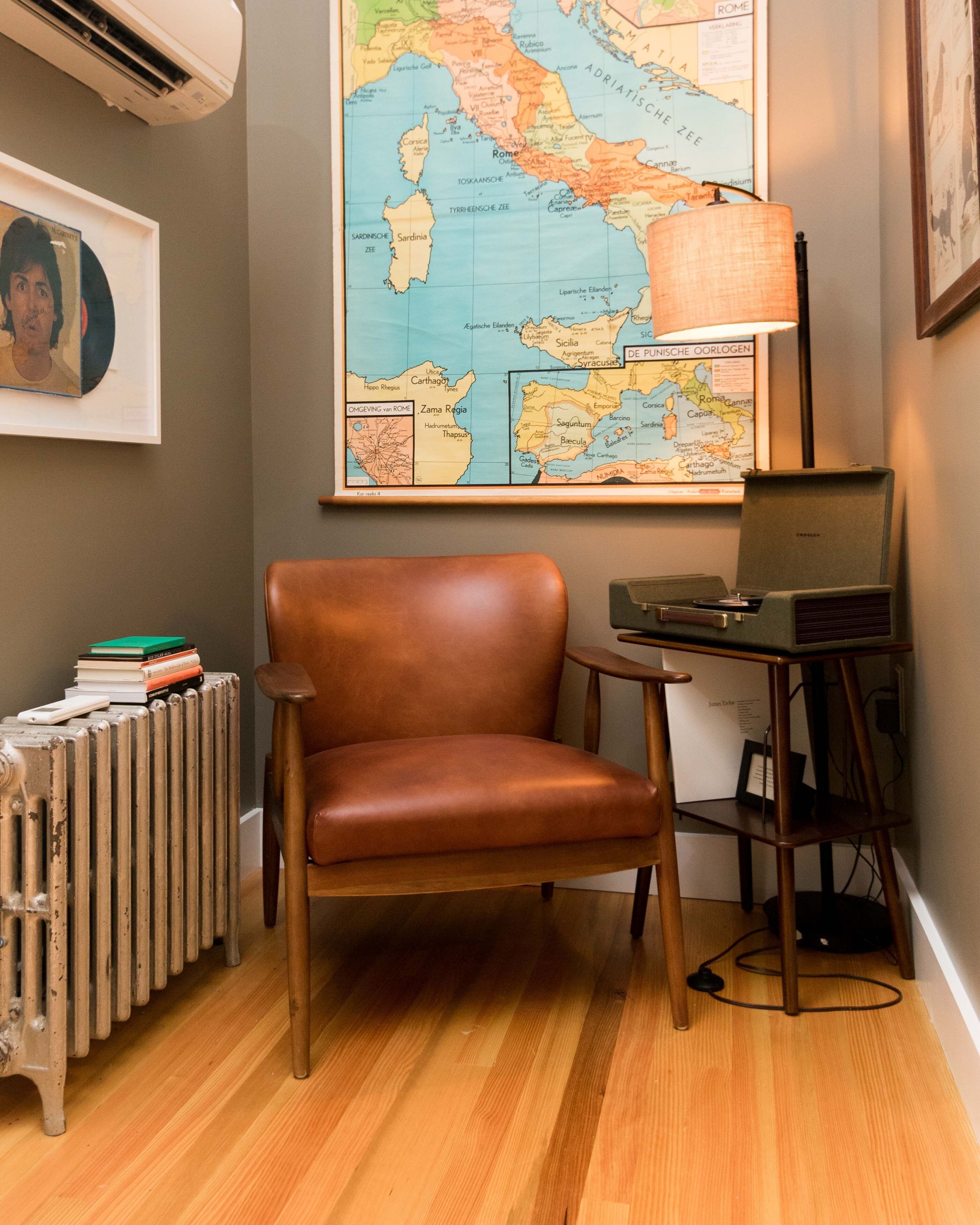 Una sala de estudio con un sillón de cuero marrón, un mapa en la pared, un radiador y un escritorio con una lámpara.