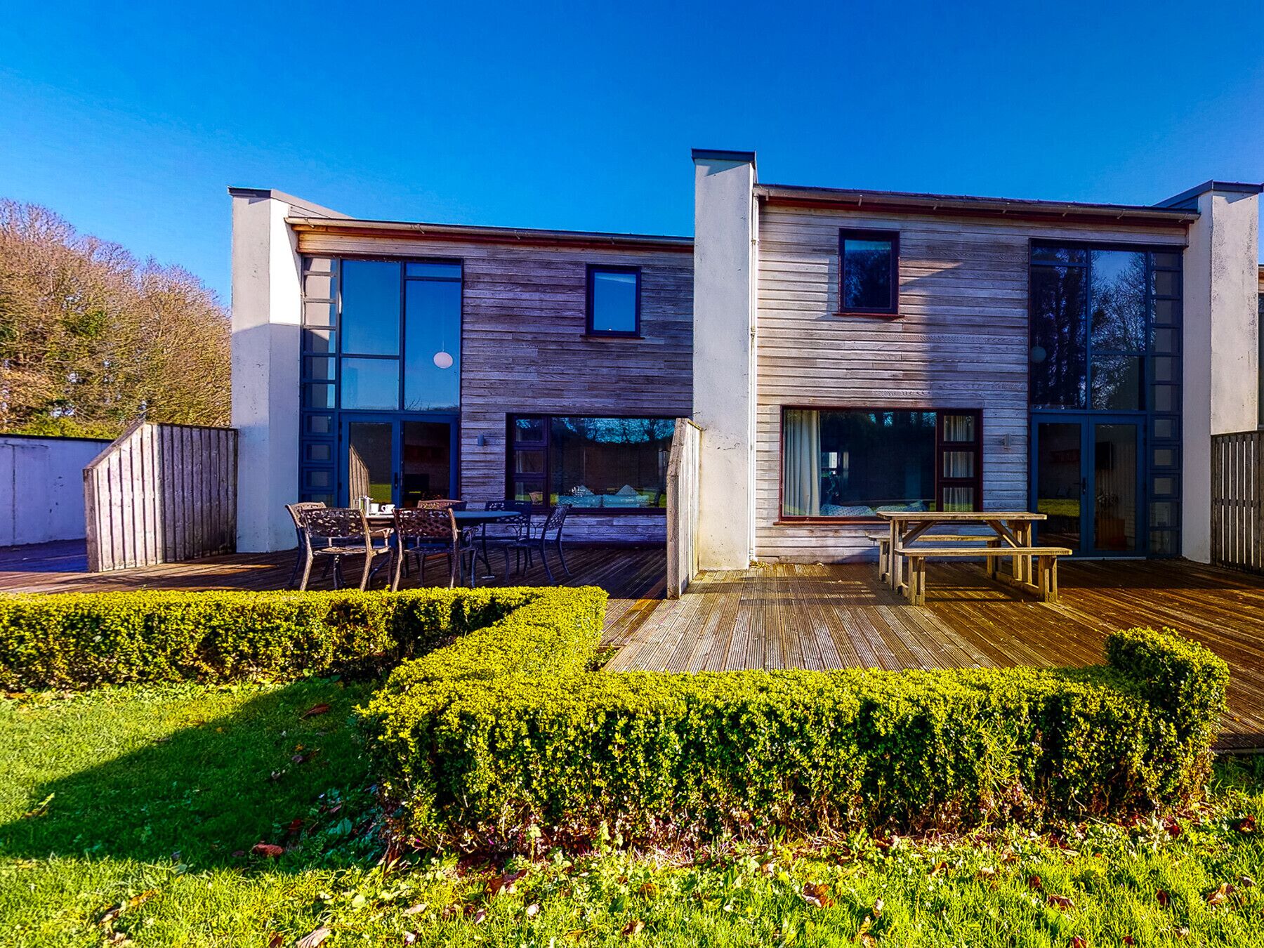 Una casa moderna de dos pisos con fachada de madera, ventanas grandes y una terraza de madera con mobiliario exterior.