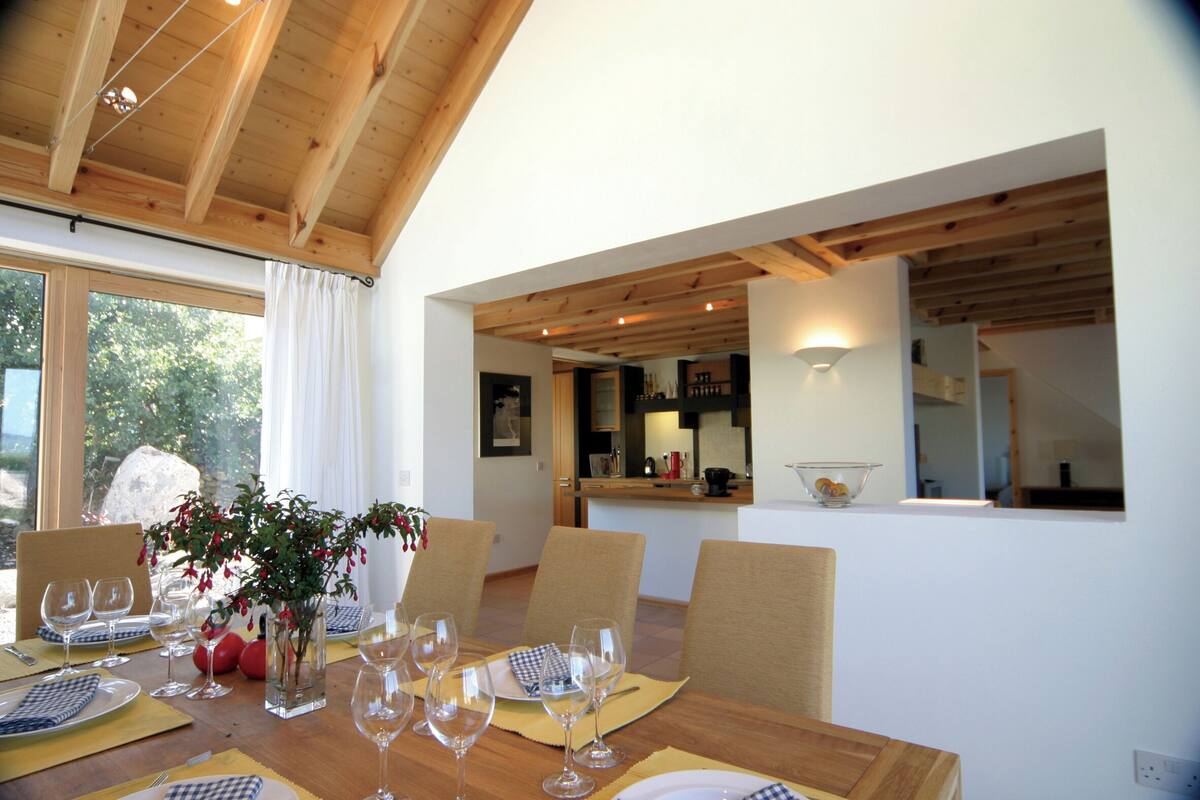 Un coin repas avec une table en bois dressée pour six personnes, un vase rempli de fleurs et une vue sur la cuisine et la salle à manger, grâce à une porte ouverte.