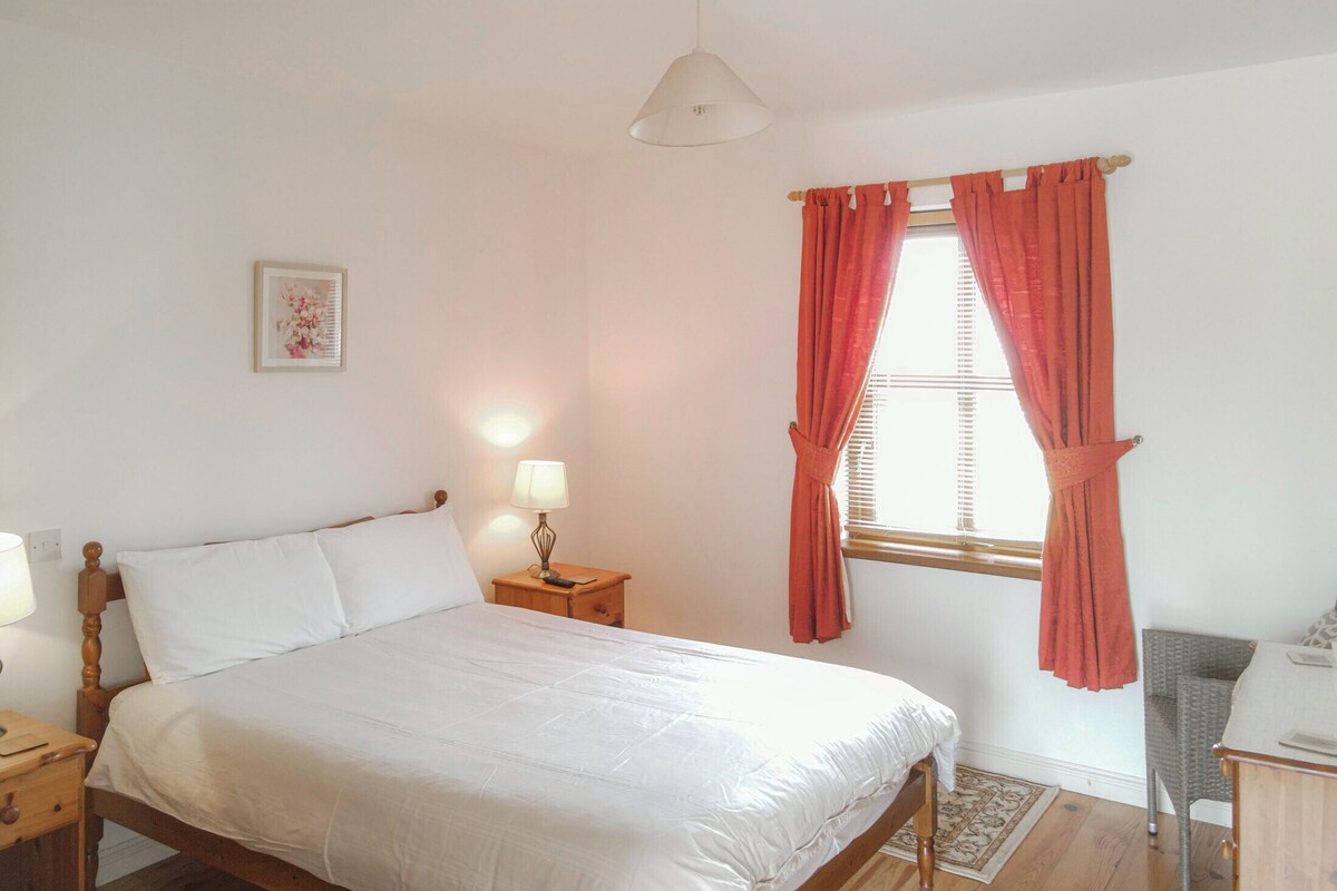 Een slaapkamer met een bed, nachtkastje, lamp, raam met gordijnen, stoel en een klein tafeltje.