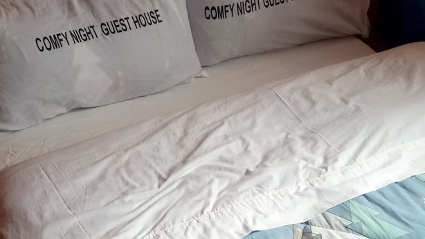 A bed with two pillows labeled "COMFY NIGHT GUEST HOUSE" and a blue quilt with a geometric pattern. - Comfy Night Guest House (Nakuru)