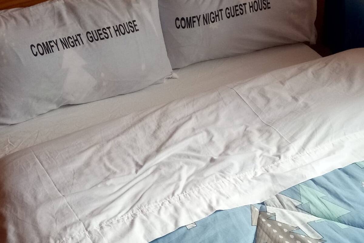 A bed with two pillows labeled "COMFY NIGHT GUEST HOUSE" and a blue quilt with a geometric pattern.