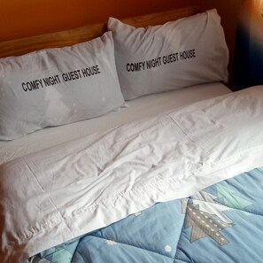 Cama com duas almofadas identificadas como "COMFY NIGHT GUEST HOUSE" e um edredom azul com padrão geométrico.