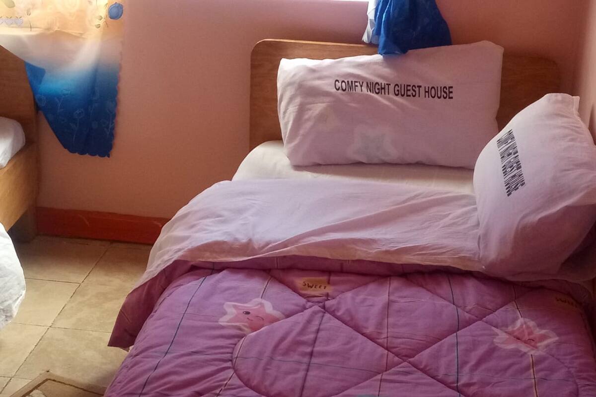A bed with a pink quilted blanket, a white pillow with "COMFY NIGHT GUEST HOUSE" text, and a blue pillow.