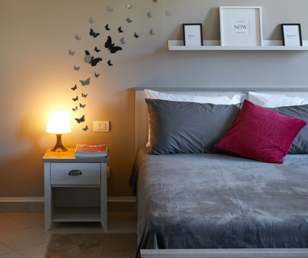 A bedroom with a bed, a nightstand with a lamp, and butterfly wall decals.
