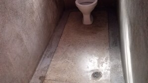 A small, narrow bathroom with a toilet, a visible pipe, and a drain. - Dakaka Lodge (Nakuru)