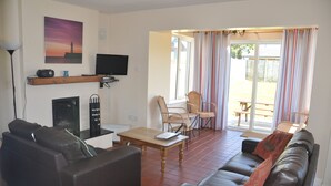 A living room with a sofa, a coffee table, a television, and a fireplace. There is a window with curtains and a view of the outside.