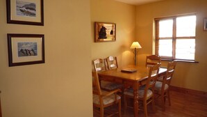 A dining area with a wooden table and chairs, framed pictures on the wall, and a window with blinds. - Fairfield Holiday Home No 13 (Dingle)