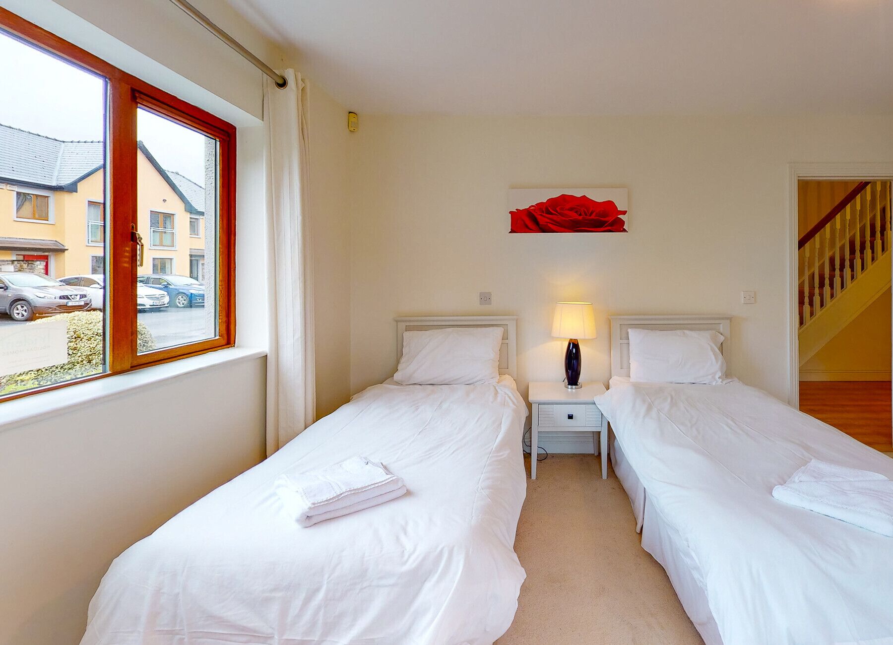 A hotel room with two single beds, a nightstand with a lamp, a red flower decoration on the wall, and a window with a view of parked cars and buildings outside.