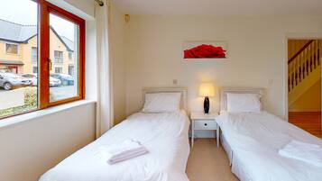 A hotel room with two single beds, a nightstand with a lamp, a red flower decoration on the wall, and a window with a view of parked cars and buildings outside.