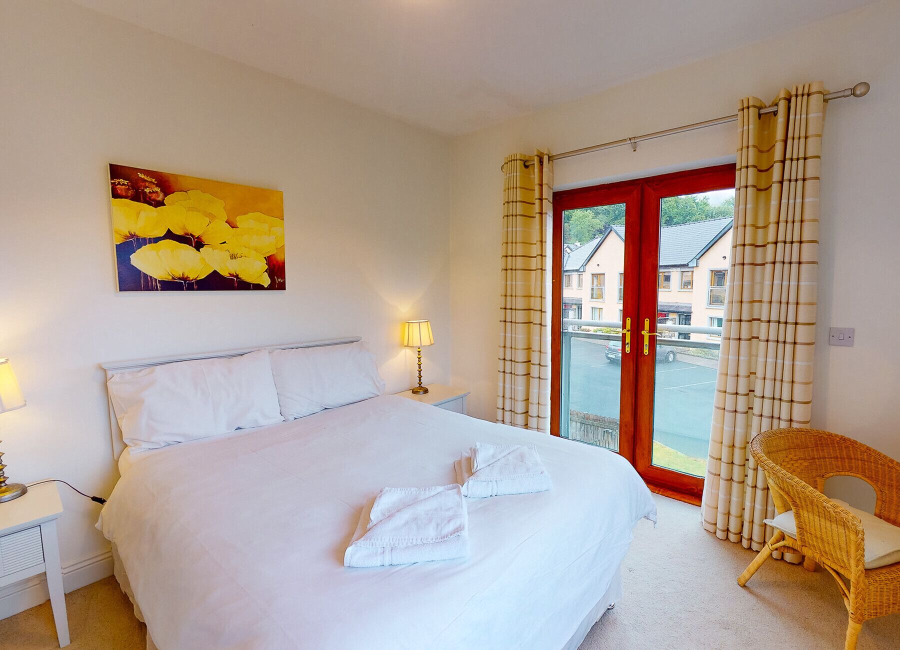 A hotel room with a bed, a nightstand with a lamp, a painting of yellow flowers, a wicker chair, and a view of buildings through a glass door.