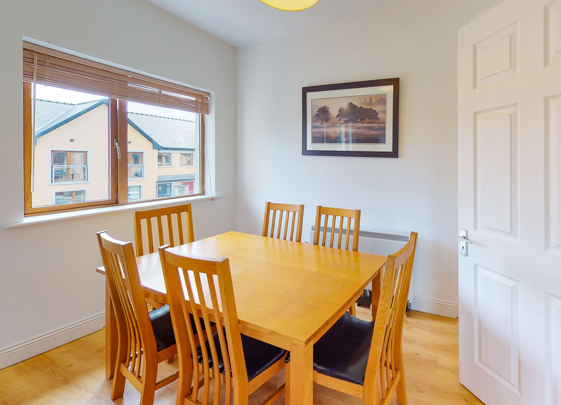 A dining room with a wooden table and chairs, a window with blinds, a framed picture on the wall, and a white door.