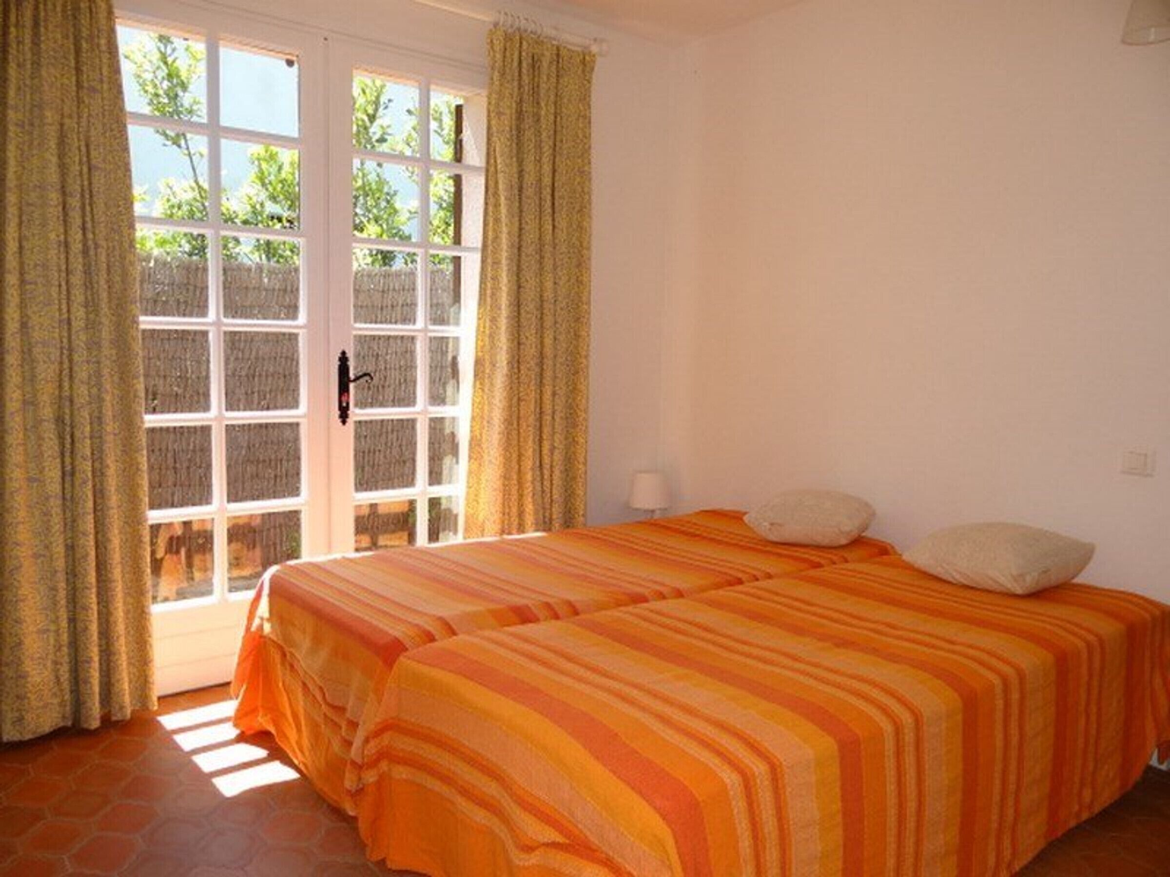 A room with two beds, a window with curtains, and a view of greenery outside.