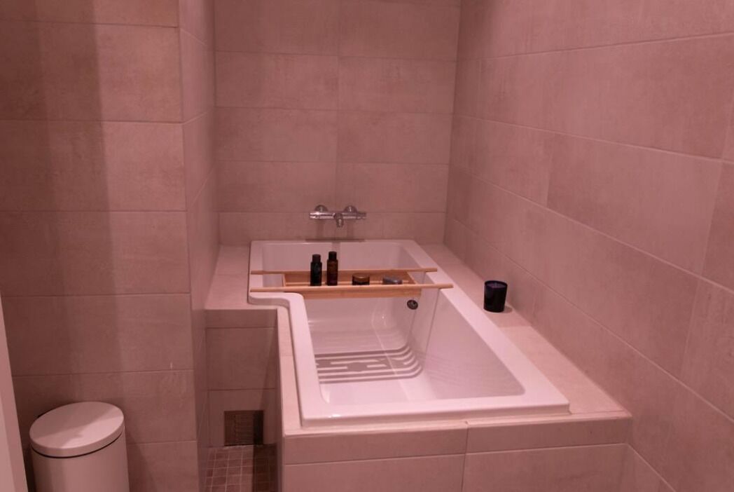 A small bathroom with a white bathtub, a shelf with toiletries, and a trash bin.