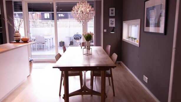 A dining area with a wooden table, chairs, a chandelier, and a view through a glass door to an outdoor seating area. - StayPlus Home near Amusement Park (Kristiansand)