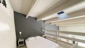 Quarto compacto com cama, mesa de cabeceira, escrivaninha e uma pequena janela.