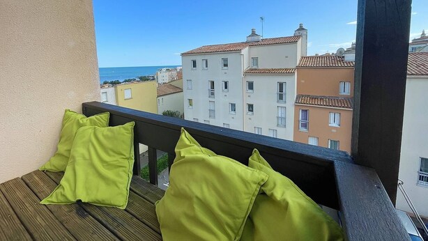 Varanda com soalho de madeira e almofadas verdes, com vista para uma paisagem urbana costeira, com edifícios e o mar.