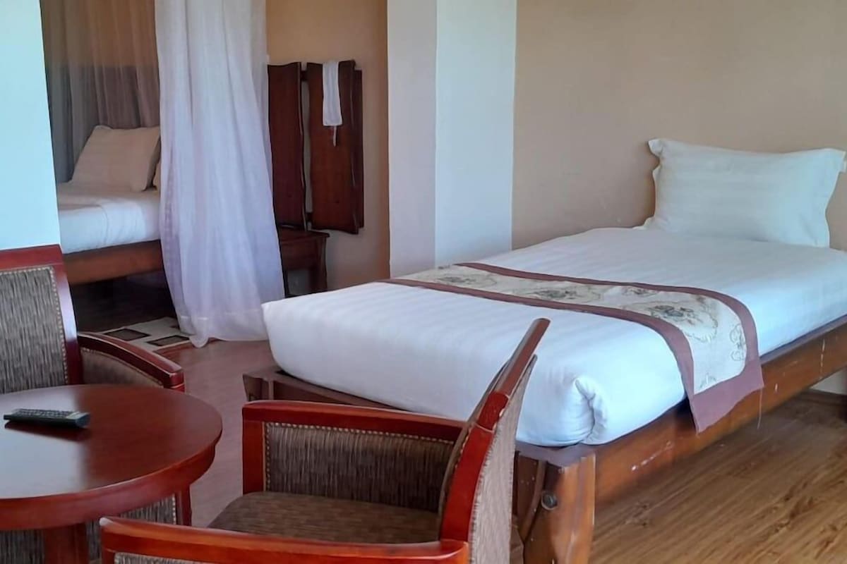A single bed with a white mattress and a brown headboard, a wooden chair, a small round table with a remote control, and a wooden wardrobe.