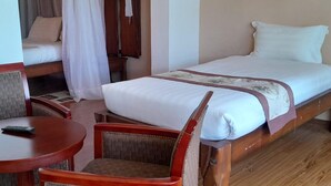 Una cama individual con cubrecama blanco y un detalle decorativo marrón. Hay una silla de madera y una mesita redonda con un control remoto encima.