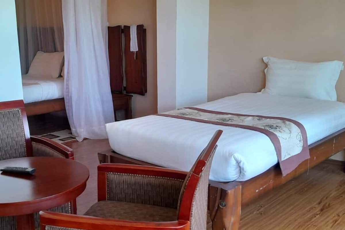 A single bed with a white bedspread and a brown decorative trim. There is a wooden chair and a small round table with a remote control on it.
