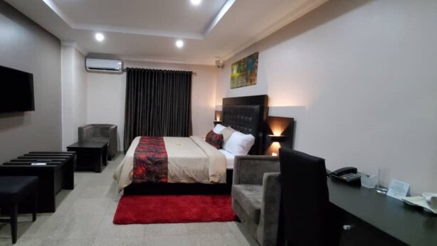 A hotel room with a bed, a desk, a chair, a TV, and an air conditioning unit. - The Lodge by Kabachi (Lagos)
