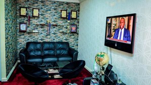 A living room with a black leather sofa, a coffee table, a globe, and a TV displaying a news broadcast. - Power Mike Guest House (Abuja)